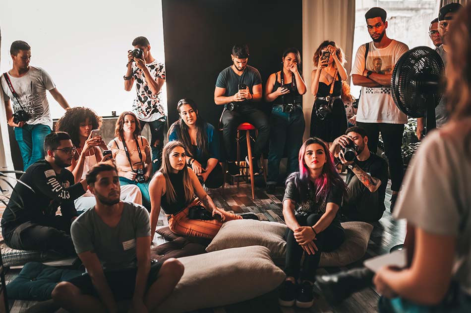 A large group of people sit on the floor in a room, looking at something out of frame