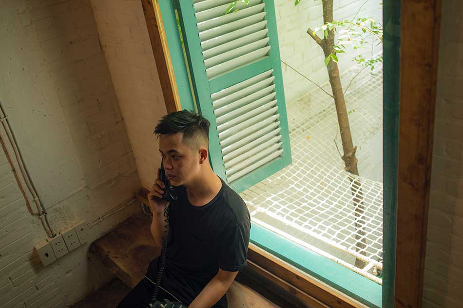 A man sits against a windowsill looking away from the camera. He is on the phone