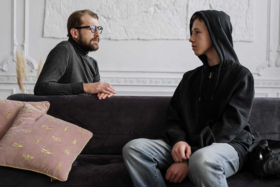A teenage boy with a black hoodie on sits on a black couch facing a bearded man wearing glasses and a turtleneck