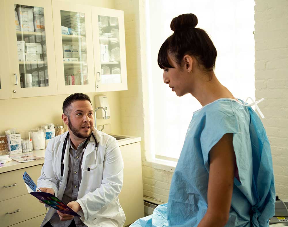 A transgender woman in a hospital gown speaking to her doctor, a transgender man, in an exam room