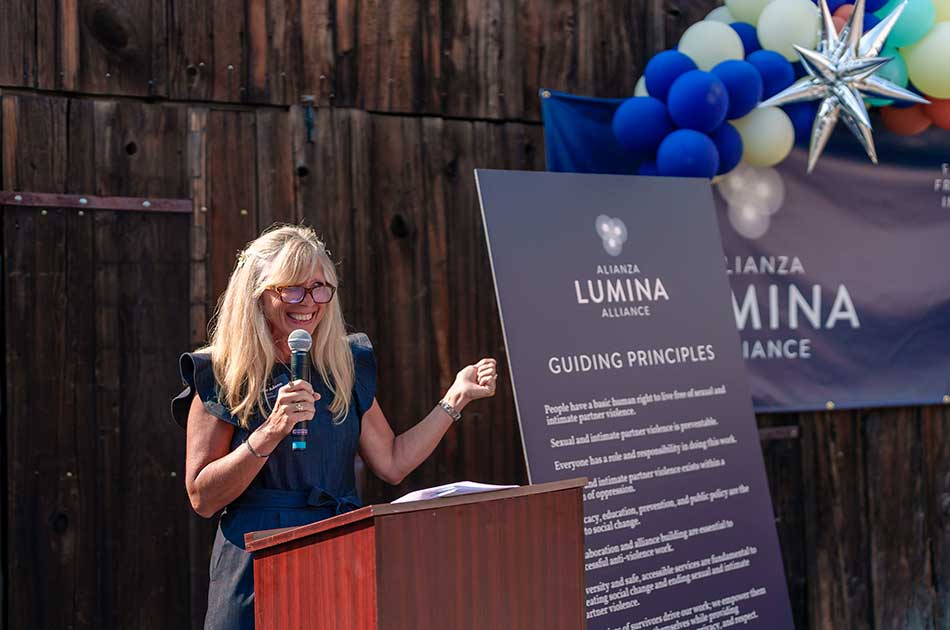 Jennifer Adams speaks into a microphone while standing next to a Lumina Alliance poster