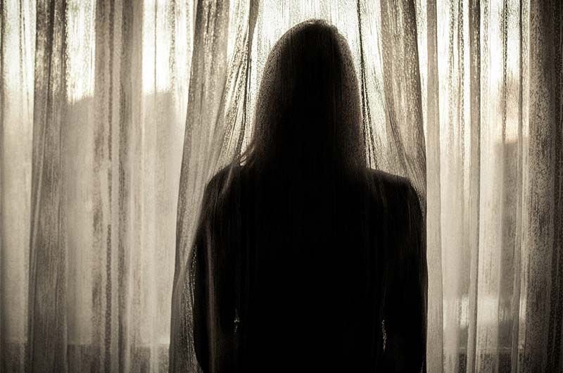Silhouette of the back of a woman looking out a window with thin drapes behind her.