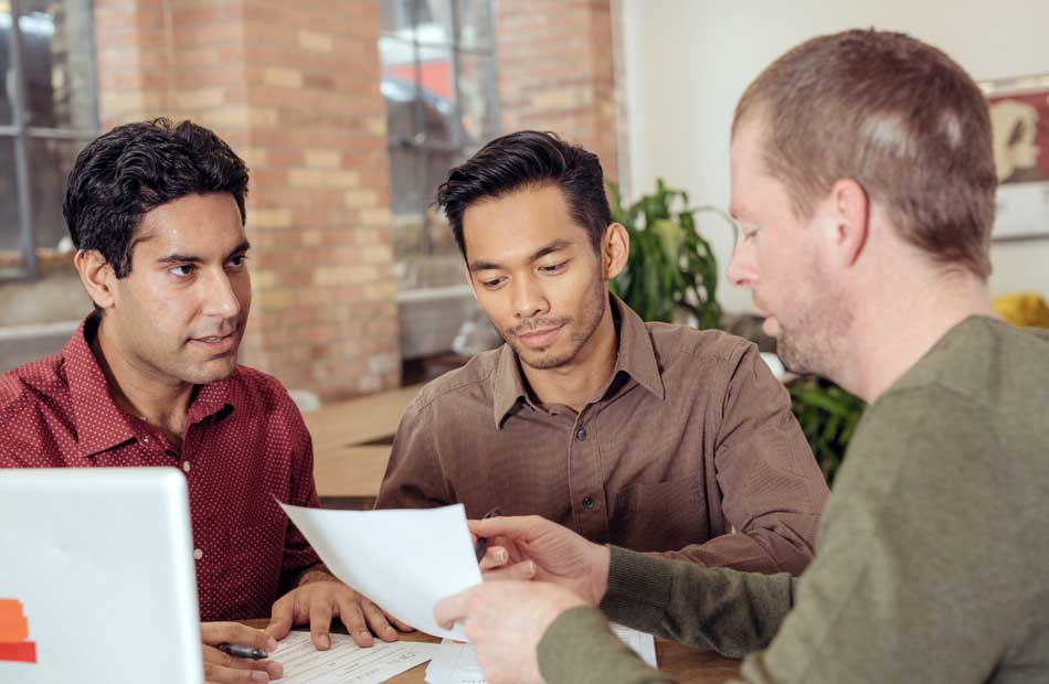 Group of three men sat at a table together discussing something.