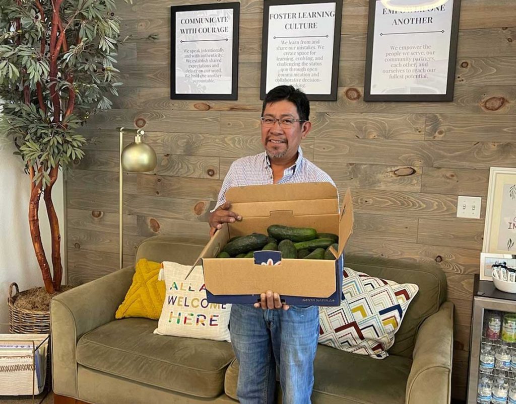 Picture of a man holding a box full of fresh cucumbers he gifted to Lumina's pantry for survivors & victims.