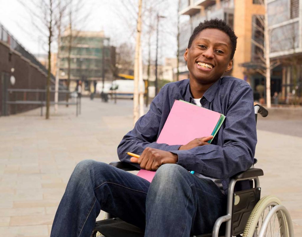 A man in a wheelchair holding folders is smiling at the camera. He is outside in a city
