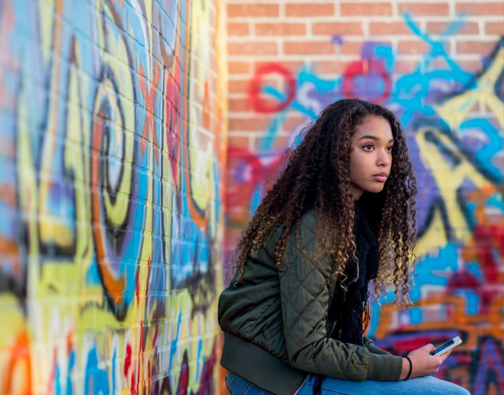 A young woman pictured in front of a graffiti-wall with her phone in one hand while looking away